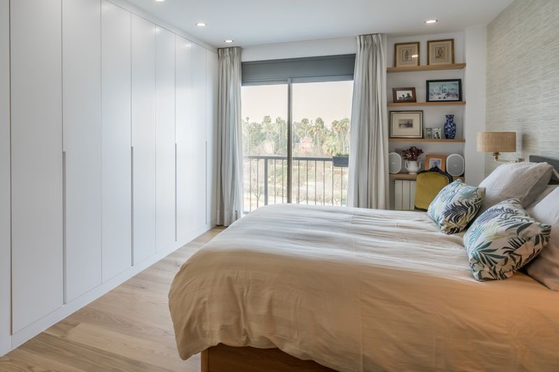 Dormitorio principal con estanteria de baldas de madera de roble acopladas a la pared y armario lacado blanco con puertas abatibles y tirador de uñero vertical